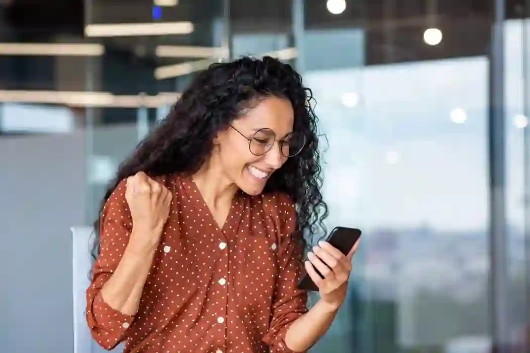 Smiling woman celebrating at the office while holding her cell phone