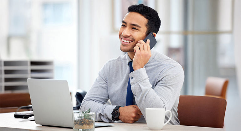 Business professional smiling during a phone call at a modern office desk with laptop, coffee mug, and potted plant, dressed in gray shirt and blue tie. Business professional smiling during a phone call at a modern office desk with laptop, coffee mug, and potted plant, dressed in gray shirt and blue tie.