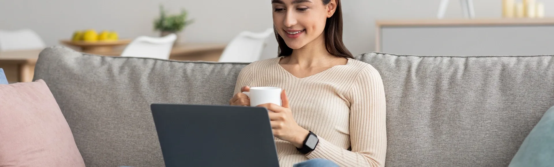 Woman sitting on a sofa, holding a cup of coffee while using a laptop at home.
