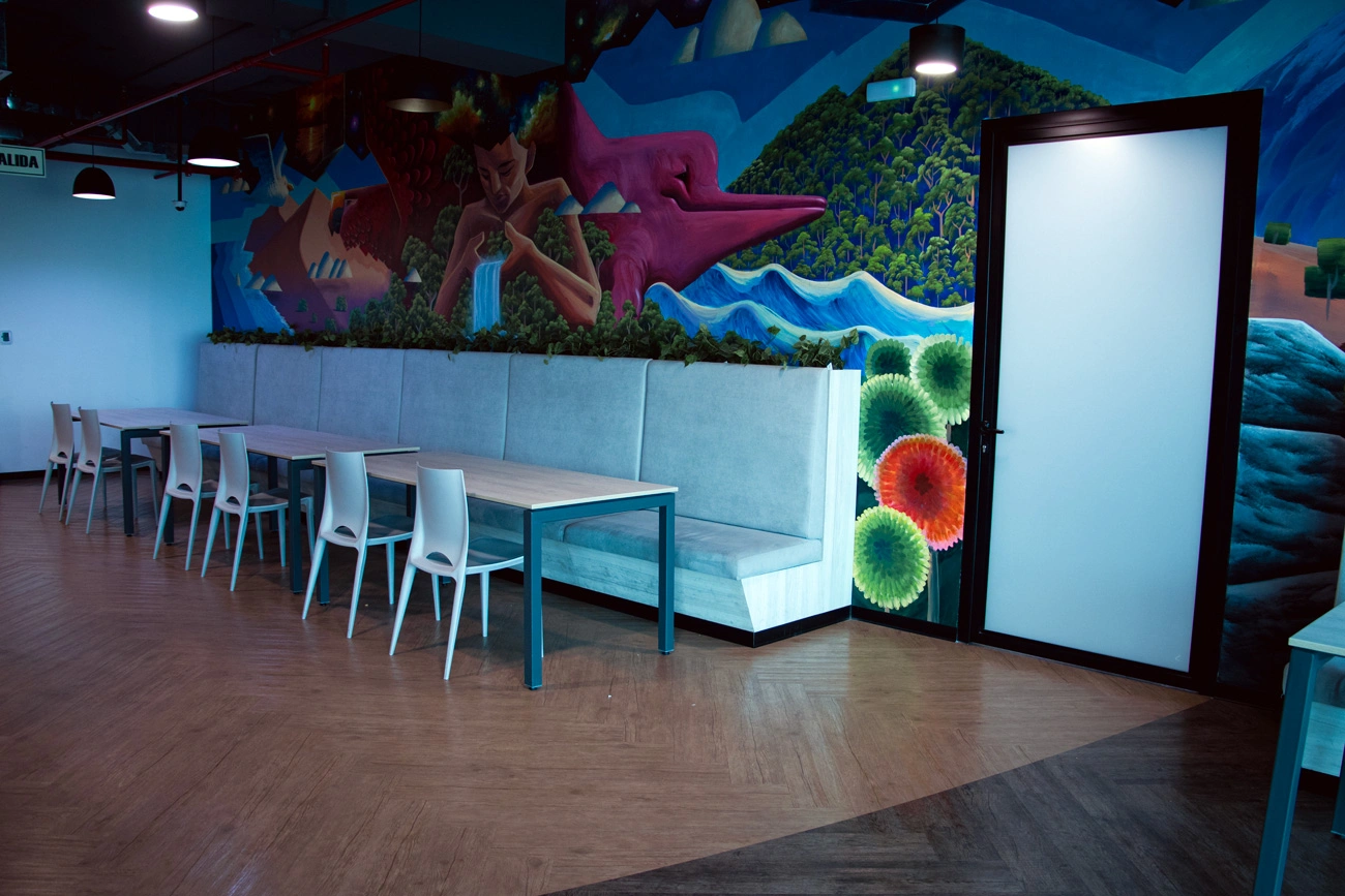 Office lounge with tables and seating beside a vibrant wall mural and open doorway.