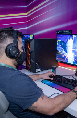 A person wearing headphones and a mask plays a game on a computer in a room with vibrant purple and pink striped walls, surrounded by gaming equipment.
