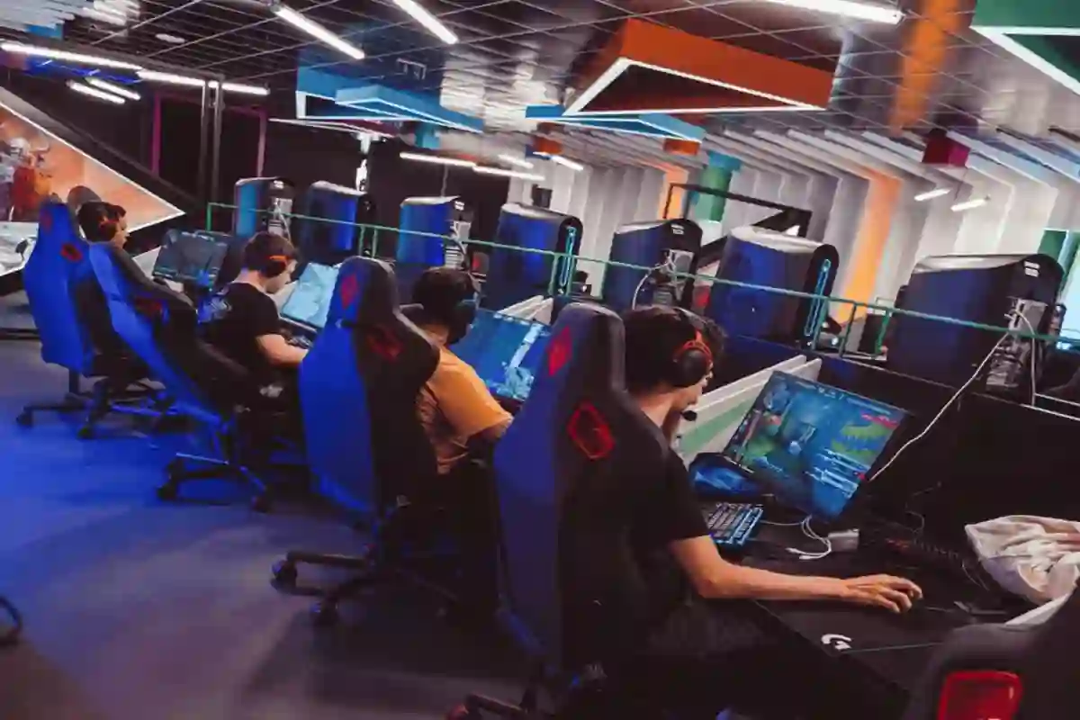 People seated at gaming stations play video games on computers, wearing headsets in a colorful, modern gaming room with neon accents and large monitors.