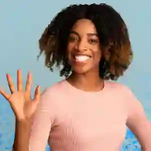 Uma pessoa sorri e acena com a mão direita, vestindo um suéter rosa. Ela está em frente a um fundo azul claro com pontos azuis espalhados abaixo.