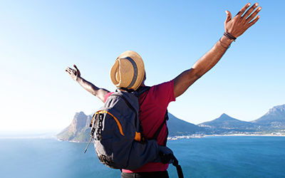 Traveler wearing a hat and backpack standing on a cliff with arms outstretched, overlooking a scenic ocean view and mountain landscape under a clear blue sky.