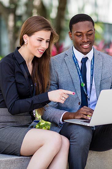 Two professionals, one in a suit and the other in business attire, sit outside, smiling at a laptop. one points at the screen while the other types, surrounded by trees.