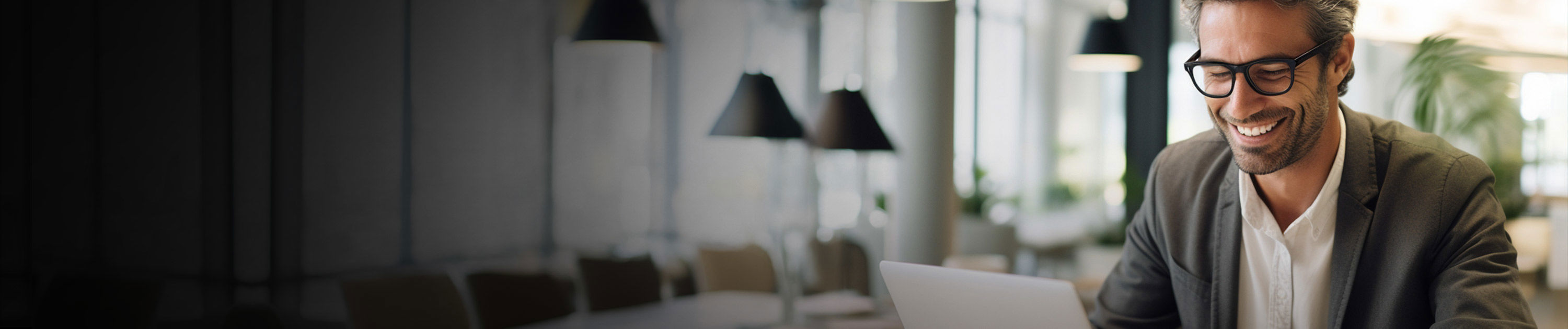a person wearing glasses and a suit smiles while using a laptop in a well-lit, modern office area with several hanging lamps and blurred background elements.