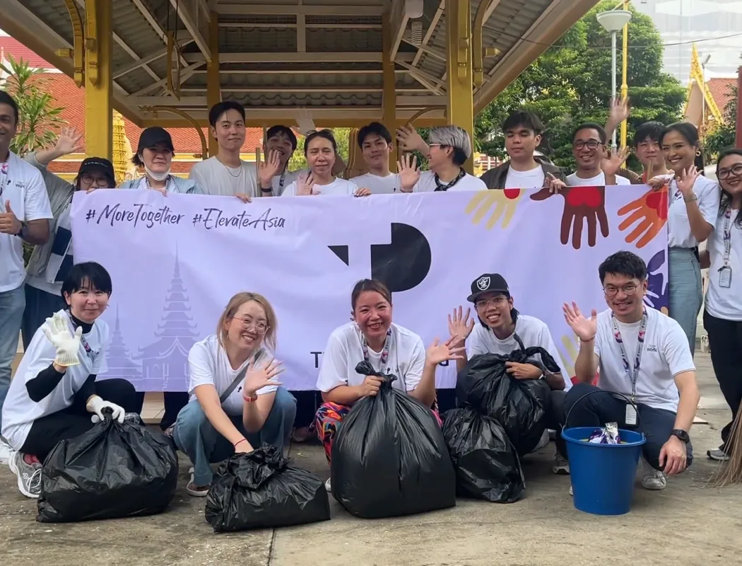 A diverse team of volunteers poses cheerfully with garbage bags, holding a "#MoreTogether #ElevateAsia" banner after a community cleanup event.