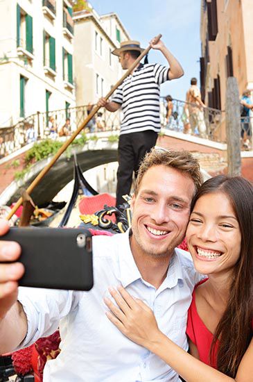 Una coppia si fa un selfie su una gondola, sorridendo da vicino. Sullo sfondo, un gondoliere rema indossando una maglietta a righe e un cappello. Sono visibili edifici e un piccolo ponte.
